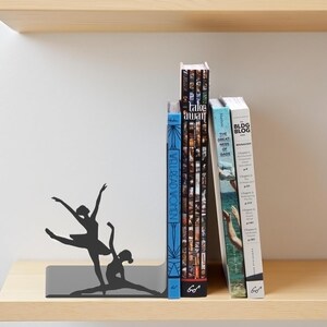 May include: A black metal bookend featuring a silhouette of two ballet dancers in mid-performance. The bookend is gray and holds several books with titles like "Take Away" and "Well Read Women" on a wooden shelf.