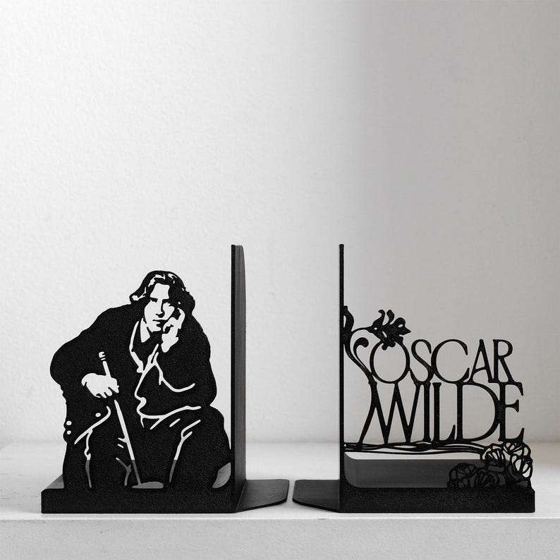 Puede incluir: Un par de sujetalibros de metal negro. Uno muestra la silueta de una persona con el pelo largo sosteniendo un bast&oacute;n, mientras que el otro presenta el texto OSCAR WILDE integrado en un dise&ntilde;o floral decorativo con l&iacute;neas curvas y hojas.