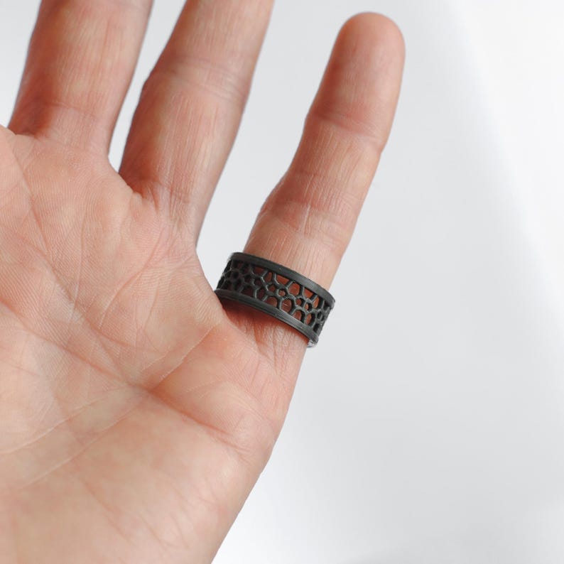 Puede incluir: Un anillo gris oscuro con un dise&ntilde;o geom&eacute;trico calado. El anillo se lleva en un dedo, mostrando su intrincado patr&oacute;n y acabado met&aacute;lico. El dise&ntilde;o del anillo presenta una serie de formas interconectadas, creando una est&eacute;tica &uacute;nica y moderna.