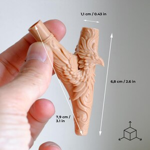 Op de afbeelding: Een beige, Y-vormig object met een gedetailleerd feniksontwerp. Het object is 7,9 cm lang, 6,8 cm hoog en 1,1 cm breed. De feniks heeft uitgespreide vleugels en is in het oppervlak gesneden. Het object wordt door een hand vastgehouden.