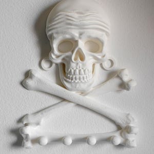 Puede incluir: Un perchero de pared blanco con forma de cr&aacute;neo humano con bandana y dos huesos cruzados debajo. El hueso inferior cuenta con cuatro peque&ntilde;os ganchos circulares para colgar objetos. Toda la estructura tiene un acabado y textura mate.