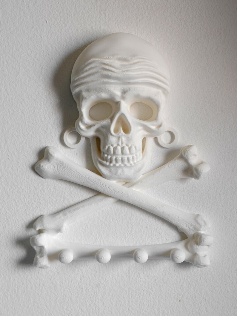 Puede incluir: Un perchero de pared blanco con forma de cr&aacute;neo humano con bandana y dos huesos cruzados debajo. El hueso inferior cuenta con cuatro peque&ntilde;os ganchos circulares para colgar objetos. Toda la estructura tiene un acabado y textura mate.