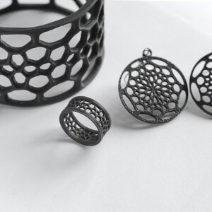 Puede incluir: Conjunto de joyas geom&eacute;tricas negras con un anillo, dos colgantes circulares y un recipiente cil&iacute;ndrico. Las piezas tienen un dise&ntilde;o abierto en forma de panal. El anillo es peque&ntilde;o y los colgantes tienen un lazo para colgar.
