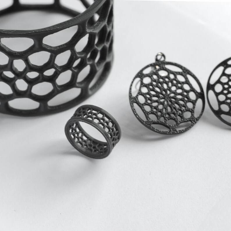 Puede incluir: Conjunto de joyas geom&eacute;tricas negras con un anillo, dos colgantes circulares y un recipiente cil&iacute;ndrico. Las piezas tienen un dise&ntilde;o abierto en forma de panal. El anillo es peque&ntilde;o y los colgantes tienen un lazo para colgar.