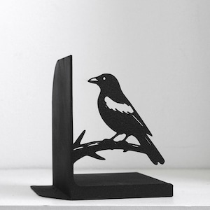 Puede incluir: Un sujetalibros de metal negro mate con la silueta de un p&aacute;jaro posado en una rama. El p&aacute;jaro tiene una mancha blanca distintiva en el ala y mira hacia la izquierda. La base en forma de L sirve de soporte para libros y tiene un acabado texturizado.