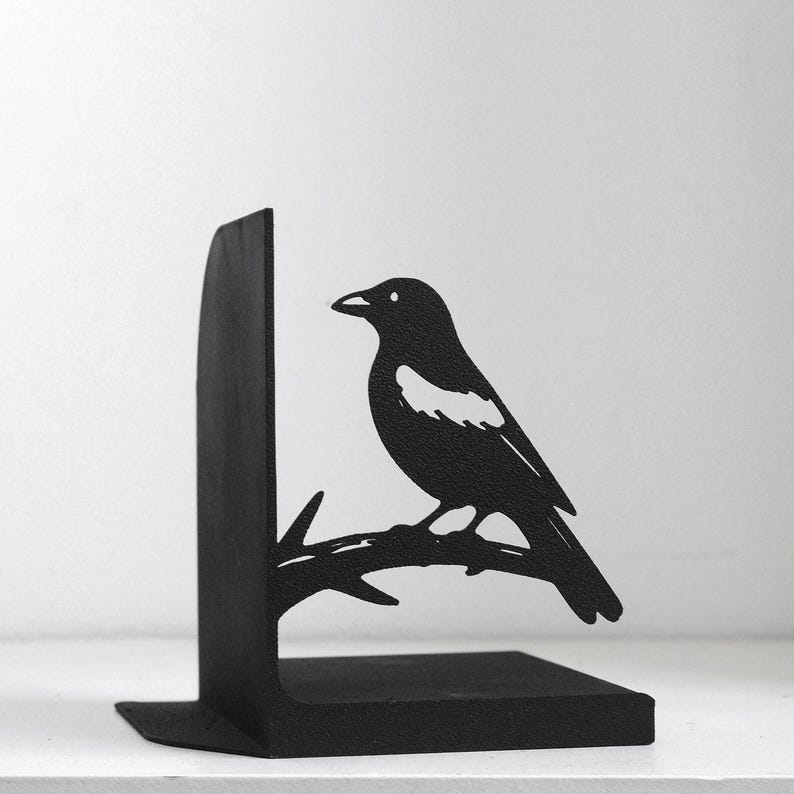 Puede incluir: Un sujetalibros de metal negro mate con la silueta de un p&aacute;jaro posado en una rama. El p&aacute;jaro tiene una mancha blanca distintiva en el ala y mira hacia la izquierda. La base en forma de L sirve de soporte para libros y tiene un acabado texturizado.