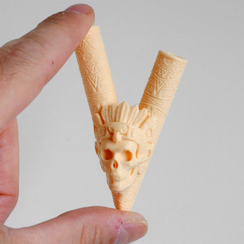 Op de afbeelding: Een lichtbeige, V-vormige rookpijp met een gedetailleerde schedel die een verentooi draagt in het midden. De twee cilindrische stelen zijn versierd met ingewikkelde geometrische patronen en het gehele object heeft een gladde, matte afwerking.