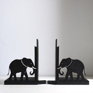 Puede incluir: Un par de sujetalibros de metal negro con la silueta de un elefante en cada pieza. El dise&ntilde;o incluye una placa de soporte vertical y una base horizontal, con la figura del elefante integrada en el lateral de cada estructura de metal resistente.
