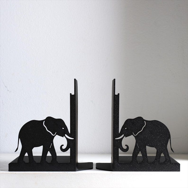 Puede incluir: Un par de sujetalibros de metal negro con la silueta de un elefante en cada pieza. El dise&ntilde;o incluye una placa de soporte vertical y una base horizontal, con la figura del elefante integrada en el lateral de cada estructura de metal resistente.