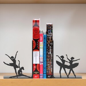May include: Black metal bookends featuring ballerina silhouettes. The bookends hold several books with colorful spines. The books include titles such as "MERINGUE GIRLS" and "WELL READ WOMEN". The bookends are on a wooden shelf.