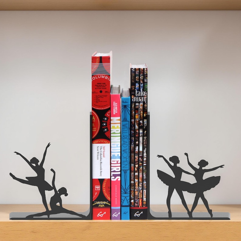 May include: Black metal bookends featuring ballerina silhouettes. The bookends hold several books with colorful spines. The books include titles such as "MERINGUE GIRLS" and "WELL READ WOMEN". The bookends are on a wooden shelf.