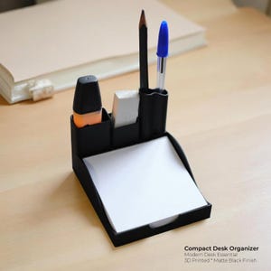 Puede incluir: Un organizador de escritorio negro con compartimentos para bolígrafos, lápices, un resaltador y una goma de borrar. También contiene un bloc de papel blanco. El texto dice "Compact Desk Organizer, Modern Desk Essential, 3D Printed * Matte Black Finish."