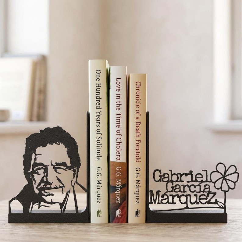 Puede incluir: Un par de sujetalibros de metal negro con un retrato en silueta de Gabriel Garc&iacute;a M&aacute;rquez en un lado y el texto Gabriel Garc&iacute;a M&aacute;rquez con un gr&aacute;fico de una flor en el otro, sosteniendo tres libros del autor en posici&oacute;n vertical.