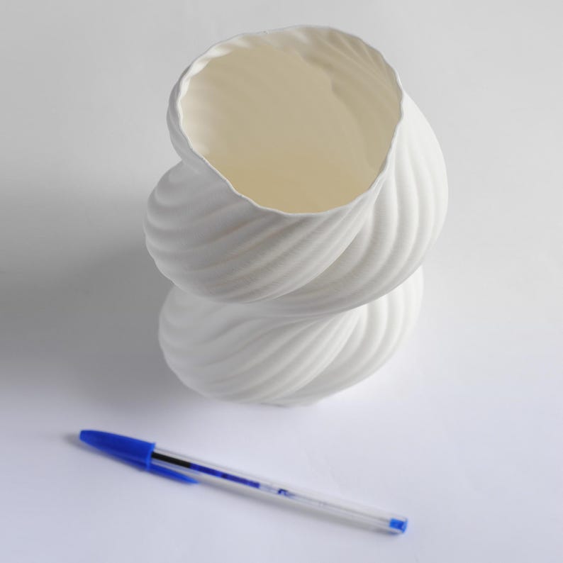 Puede incluir: Un jarr&oacute;n decorativo blanco en forma de espiral con una superficie texturizada. El jarr&oacute;n tiene una gran abertura en la parte superior y otra m&aacute;s peque&ntilde;a en la inferior. Un bol&iacute;grafo azul est&aacute; al lado del jarr&oacute;n.