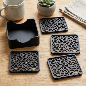 Pode incluir: Um conjunto de quatro bases para copos quadradas pretas com um intrincado padrão de grelha geométrica vazada, acompanhado por um suporte quadrado preto a condizer com um design recortado em cada lado para fácil acesso.