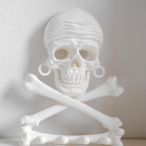 Puede incluir: Un perchero de pared blanco con forma de calavera que lleva un pa&ntilde;uelo y pendientes de aro, situado sobre dos huesos cruzados. La base consiste en un hueso horizontal con cuatro ganchos redondeados para colgar diversos objetos.