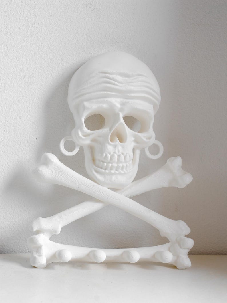 Puede incluir: Un perchero de pared blanco con forma de calavera que lleva un pa&ntilde;uelo y pendientes de aro, situado sobre dos huesos cruzados. La base consiste en un hueso horizontal con cuatro ganchos redondeados para colgar diversos objetos.
