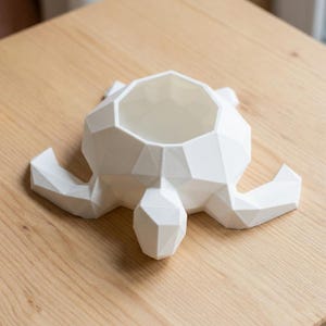 Puede incluir: Una maceta blanca con forma de tortuga geom&eacute;trica. La maceta tiene un cuenco grande y abierto para plantas y cuatro patas cortas. La tortuga est&aacute; hecha de un material duro y liso y se encuentra sobre una superficie de madera clara.