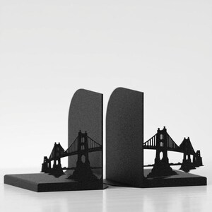 May include: A pair of black metal bookends featuring a laser-cut silhouette of a suspension bridge. Each L-shaped piece has a vertical panel displaying the bridge structure and a flat base to support books, finished with a matte black coating.