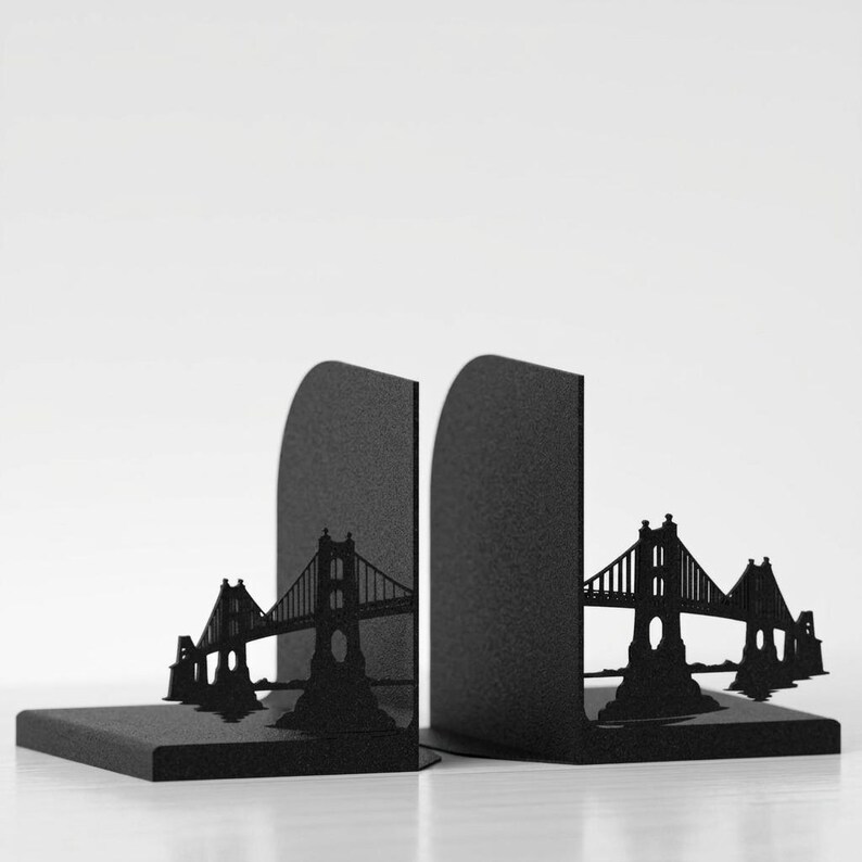 May include: A pair of black metal bookends featuring a laser-cut silhouette of a suspension bridge. Each L-shaped piece has a vertical panel displaying the bridge structure and a flat base to support books, finished with a matte black coating.