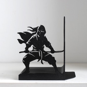Puede incluir: Un sujetalibros de metal negro con la silueta de un ninja en una postura din&aacute;mica. La figura se muestra en cuclillas con una espada sostenida horizontalmente, vistiendo un atuendo tradicional con una capa que fluye sobre un soporte vertical.