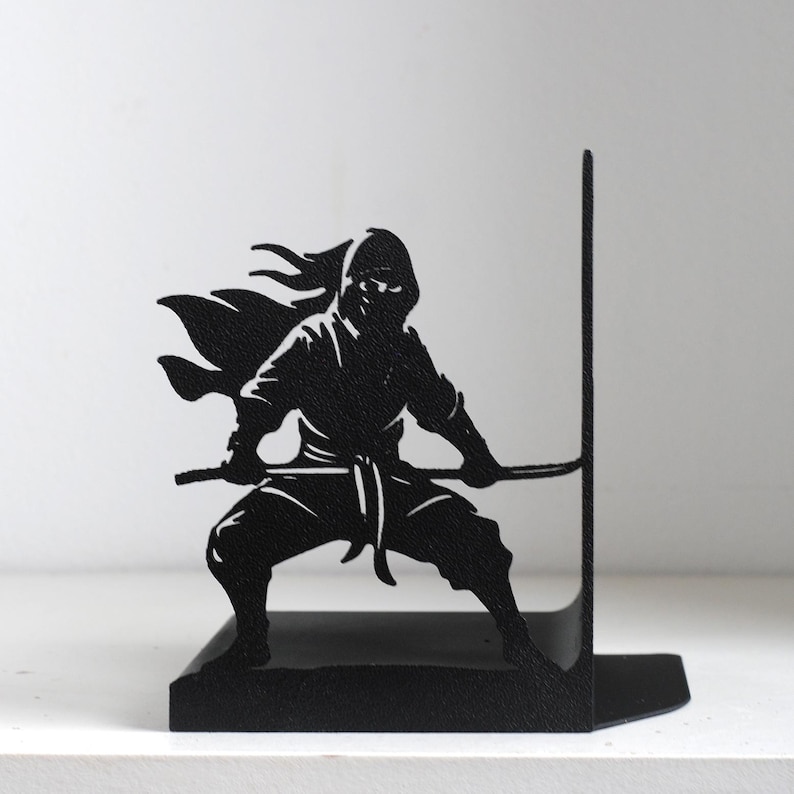 Puede incluir: Un sujetalibros de metal negro con la silueta de un ninja en una postura din&aacute;mica. La figura se muestra en cuclillas con una espada sostenida horizontalmente, vistiendo un atuendo tradicional con una capa que fluye sobre un soporte vertical.