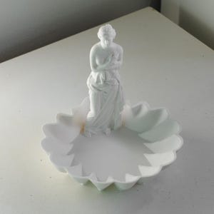 Puede incluir: Una bandeja decorativa de color blanco mate que presenta una figura central de pie envuelta en tela. La bandeja tiene un dise&ntilde;o de borde festoneado similar a p&eacute;talos. Todo el objeto es de color blanco monocrom&aacute;tico con un acabado liso.