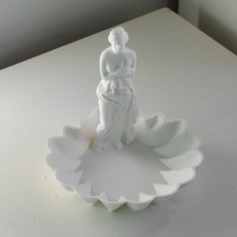 Puede incluir: Una bandeja decorativa de color blanco mate que presenta una figura central de pie envuelta en tela. La bandeja tiene un dise&ntilde;o de borde festoneado similar a p&eacute;talos. Todo el objeto es de color blanco monocrom&aacute;tico con un acabado liso.