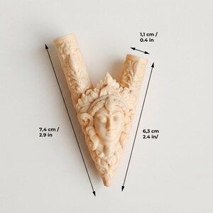 Puede incluir: Un objeto tallado en forma de V de color beige que presenta un rostro humano central con tocado ornamentado y motivos de hojas. El objeto mide 7,4 cm de largo total, 6,3 cm en el lado derecho y 1,1 cm de ancho en la abertura superior.
