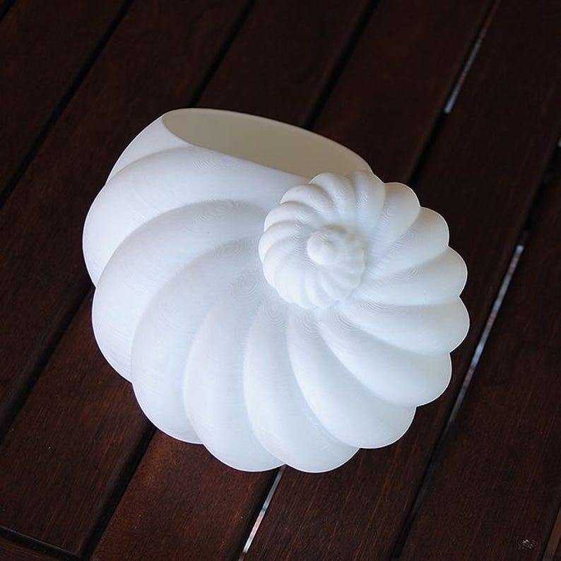 May include: A white, nautilus-shaped decorative object. The spiral design features a central, rounded element and a series of curved, overlapping sections. The object is made of a smooth, matte material and sits on a dark brown surface.