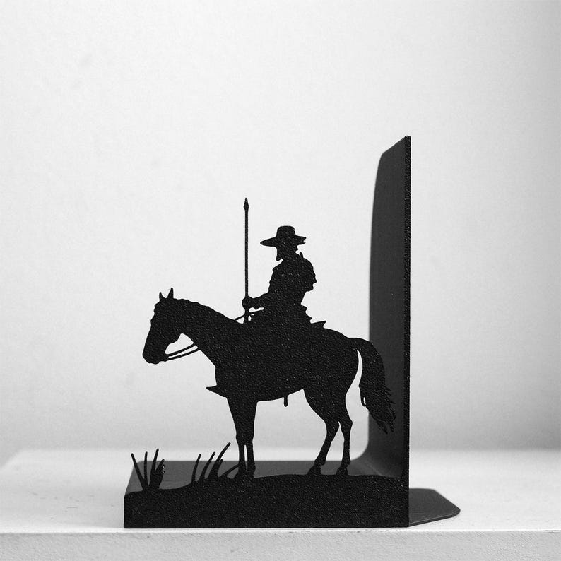 Puede incluir: Un sujetalibros de metal negro con la silueta de un jinete a caballo sosteniendo una lanza larga. El dise&ntilde;o incluye una base plana y un panel de soporte vertical, con peque&ntilde;os detalles en forma de hierba a los pies del caballo.