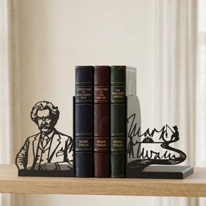 Puede incluir: Un juego de sujetalibros de metal negro con una silueta de Mark Twain en la pieza izquierda y una inscripci&oacute;n decorativa Mark Twain con una escena de r&iacute;o y una figura pescando en la pieza derecha, sosteniendo tres libros antiguos.
