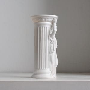 Puede incluir: Un florero cil&iacute;ndrico de color blanco mate con un dise&ntilde;o de columna estriada. Un lado del florero est&aacute; adornado con una figura esculpida con ropa drapeada, con los brazos levantados para sostener el borde superior de la columna. Acabado liso.