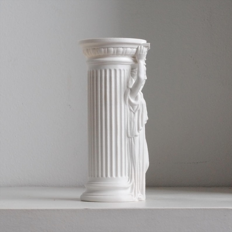 Puede incluir: Un florero cil&iacute;ndrico de color blanco mate con un dise&ntilde;o de columna estriada. Un lado del florero est&aacute; adornado con una figura esculpida con ropa drapeada, con los brazos levantados para sostener el borde superior de la columna. Acabado liso.