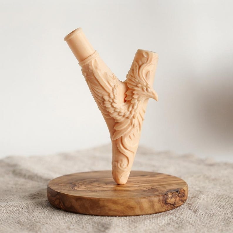 Puede incluir: Una pipa de fumar en forma de Y de color beige claro, tallada con un dise&ntilde;o detallado de f&eacute;nix, con un acabado liso y una boquilla cil&iacute;ndrica, montada verticalmente sobre una base circular de madera pulida con vetas naturales.