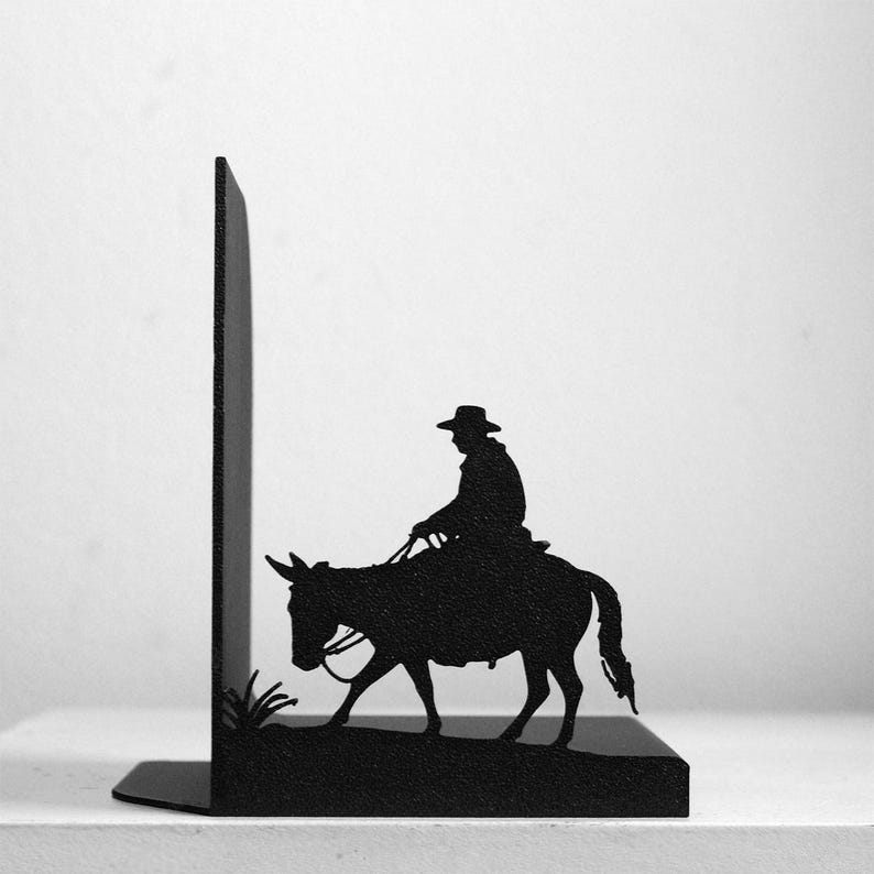 Puede incluir: Un sujetalibros de metal negro con la silueta de una persona con sombrero de ala ancha montada en una mula. El dise&ntilde;o incluye un peque&ntilde;o parche de hierba en la base y un panel vertical que sirve de soporte para los libros en una estanter&iacute;a.