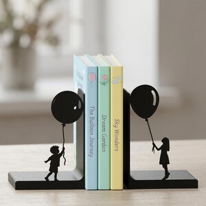 May include: A pair of black metal bookends featuring silhouettes of a child holding a balloon. The left bookend shows a child looking up at a balloon, and the right shows a child standing with a balloon. Three books are held between them.