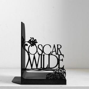 Puede incluir: Un sujetalibros de metal negro con un dise&ntilde;o recortado del nombre Oscar Wilde en una fuente estilizada. La estructura met&aacute;lica incluye elementos florales decorativos integrados en las letras y una base s&oacute;lida en forma de L.