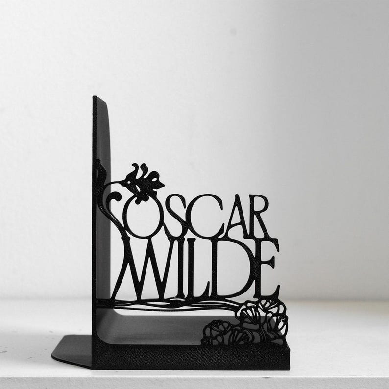 Puede incluir: Un sujetalibros de metal negro con un dise&ntilde;o recortado del nombre Oscar Wilde en una fuente estilizada. La estructura met&aacute;lica incluye elementos florales decorativos integrados en las letras y una base s&oacute;lida en forma de L.
