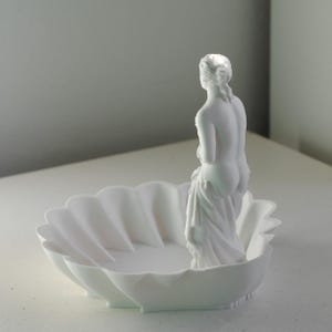 Puede incluir: Un cuenco decorativo de acabado blanco mate con forma de concha festoneada. Una peque&ntilde;a y detallada figura blanca de una persona est&aacute; de pie dentro del cuenco, mirando hacia el lado opuesto al espectador. El objeto es monocromo y liso.