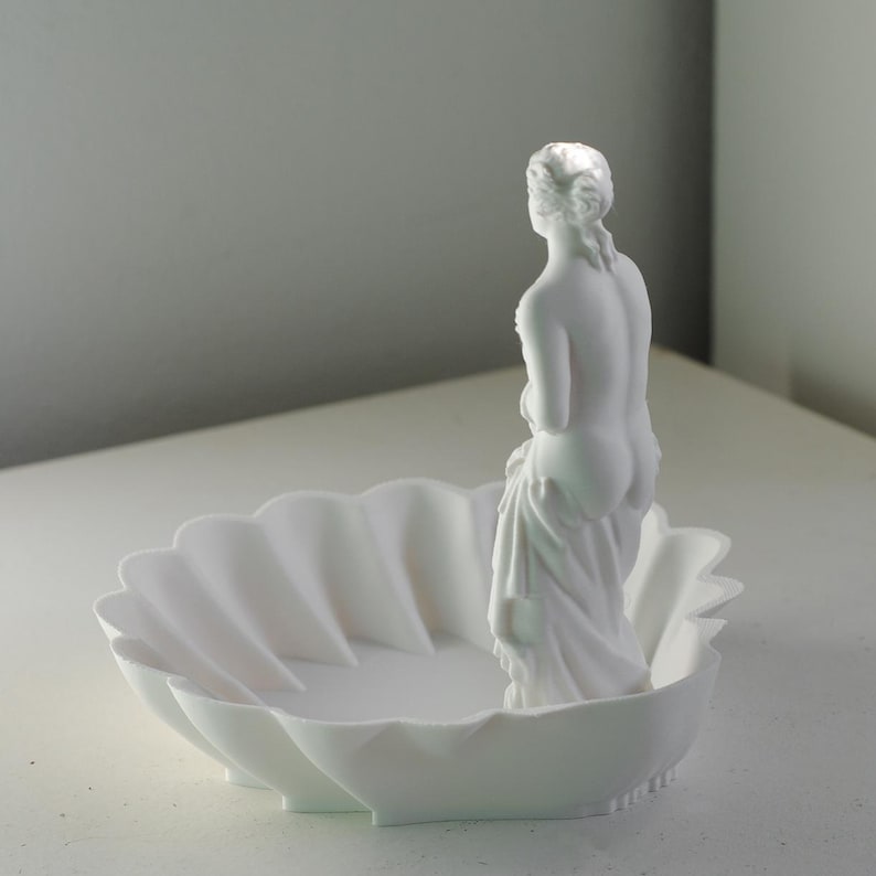 Puede incluir: Un cuenco decorativo de acabado blanco mate con forma de concha festoneada. Una peque&ntilde;a y detallada figura blanca de una persona est&aacute; de pie dentro del cuenco, mirando hacia el lado opuesto al espectador. El objeto es monocromo y liso.