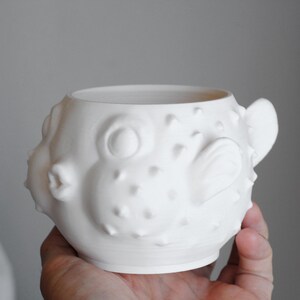 Puede incluir: Una taza de cer&aacute;mica blanca mate con forma de pez globo. La taza presenta un cuerpo redondeado con p&uacute;as puntiagudas en relieve por toda la superficie, una boca prominente, grandes ojos circulares y una peque&ntilde;a aleta en el lateral.