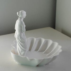 Puede incluir: Un cuenco decorativo de color blanco mate con forma de concha festoneada, que presenta una figura esculpida de una estatua cl&aacute;sica de pie en un lado del interior. El objeto es totalmente monocrom&aacute;tico con un acabado liso y uniforme.