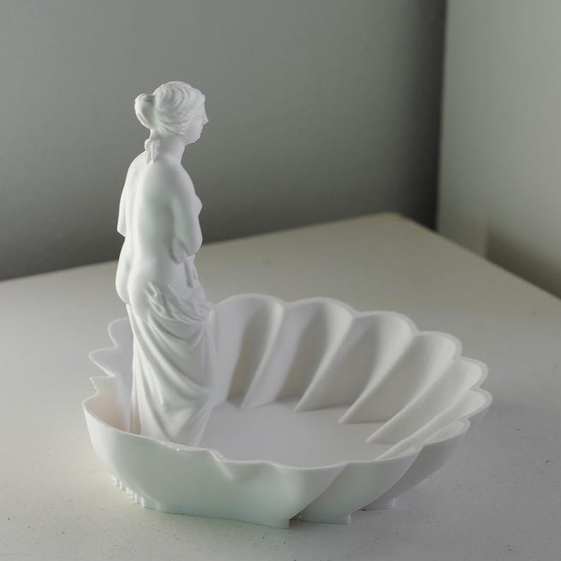 Puede incluir: Un cuenco decorativo de color blanco mate con forma de concha festoneada, que presenta una figura esculpida de una estatua cl&aacute;sica de pie en un lado del interior. El objeto es totalmente monocrom&aacute;tico con un acabado liso y uniforme.