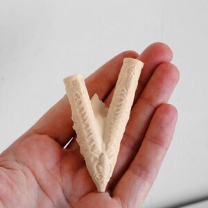Puede incluir: Un componente en forma de V, de color beige claro, hecho de pl&aacute;stico o resina moldeada, con intrincados patrones de relieve floral y volutas a lo largo de sus dos brazos. El objeto tiene una abertura cil&iacute;ndrica hueca en cada extremo.