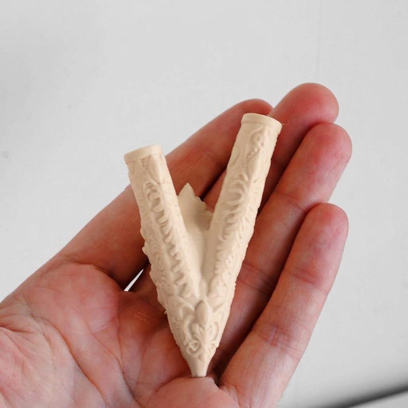 Puede incluir: Un componente en forma de V, de color beige claro, hecho de pl&aacute;stico o resina moldeada, con intrincados patrones de relieve floral y volutas a lo largo de sus dos brazos. El objeto tiene una abertura cil&iacute;ndrica hueca en cada extremo.
