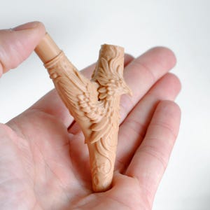 Op de afbeelding: Een beige, Y-vormige sculptuur met gedetailleerde gravures van een vogel met uitgespreide vleugels. De sculptuur wordt in een hand gehouden tegen een witte achtergrond. De veren van de vogel en de steel zijn ingewikkeld ontworpen.