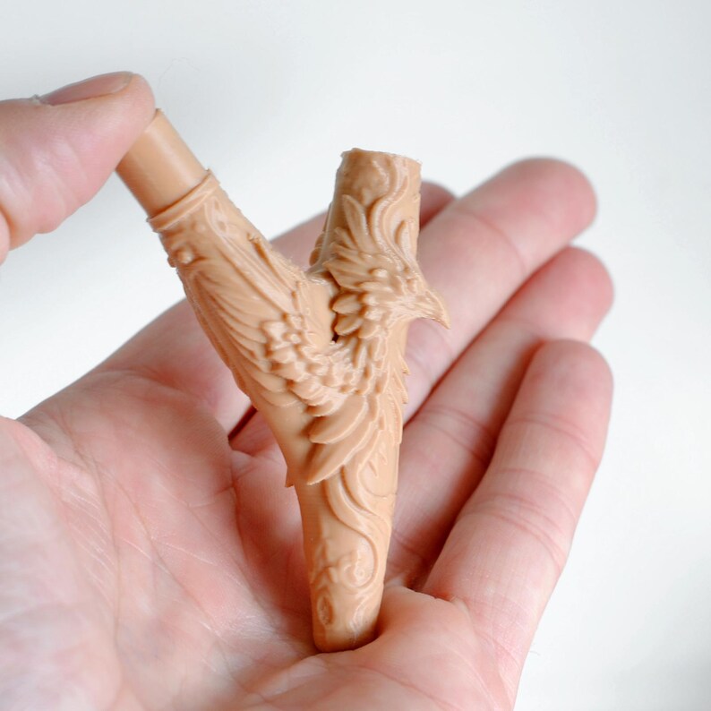 Op de afbeelding: Een beige, Y-vormige sculptuur met gedetailleerde gravures van een vogel met uitgespreide vleugels. De sculptuur wordt in een hand gehouden tegen een witte achtergrond. De veren van de vogel en de steel zijn ingewikkeld ontworpen.