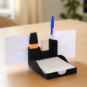Puede incluir: Un organizador de escritorio negro con compartimentos para bol&iacute;grafos, un resaltador y una goma de borrar. Contiene una pila de papel de notas blanco y dos notas que dicen "CALL OFFICE" y "BUY MILK". Se ve un bol&iacute;grafo azul.
