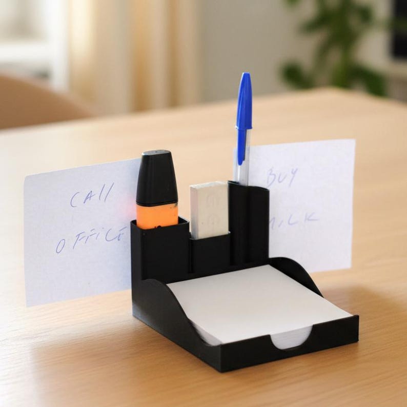 Puede incluir: Un organizador de escritorio negro con compartimentos para bol&iacute;grafos, un resaltador y una goma de borrar. Contiene una pila de papel de notas blanco y dos notas que dicen "CALL OFFICE" y "BUY MILK". Se ve un bol&iacute;grafo azul.